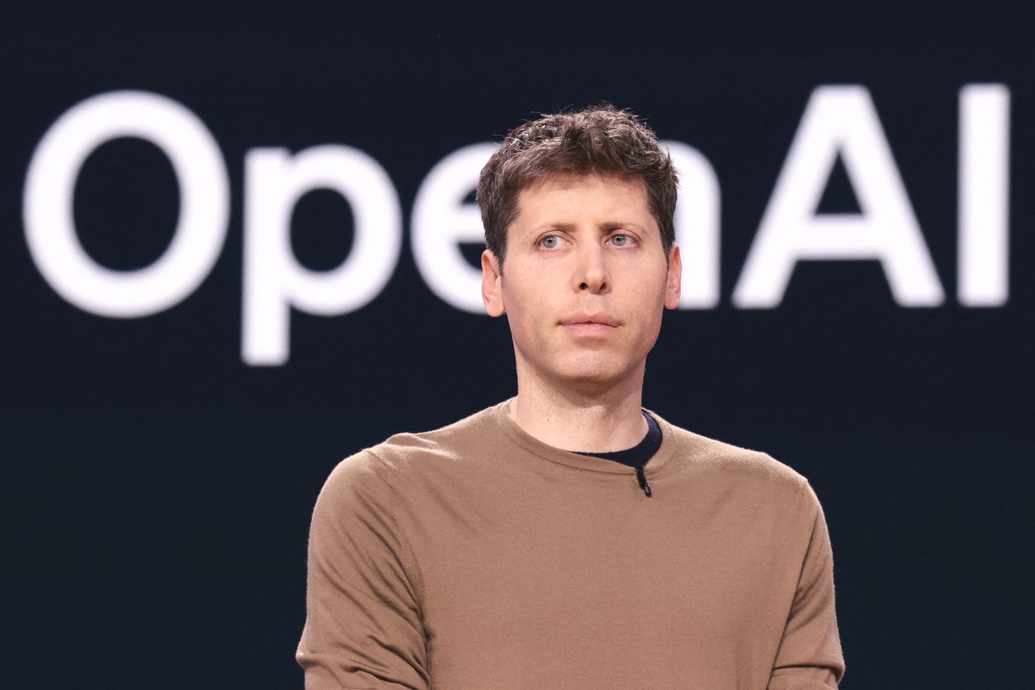 OpenAI CEO Sam Altman on stage in front of a large OpenAI sign. The mixed messaging from Altman, oscillating between 'augment workers' and 'AI will replace the CEO,' has been a major contributor to public confusion about what the technology will actually do to jobs.